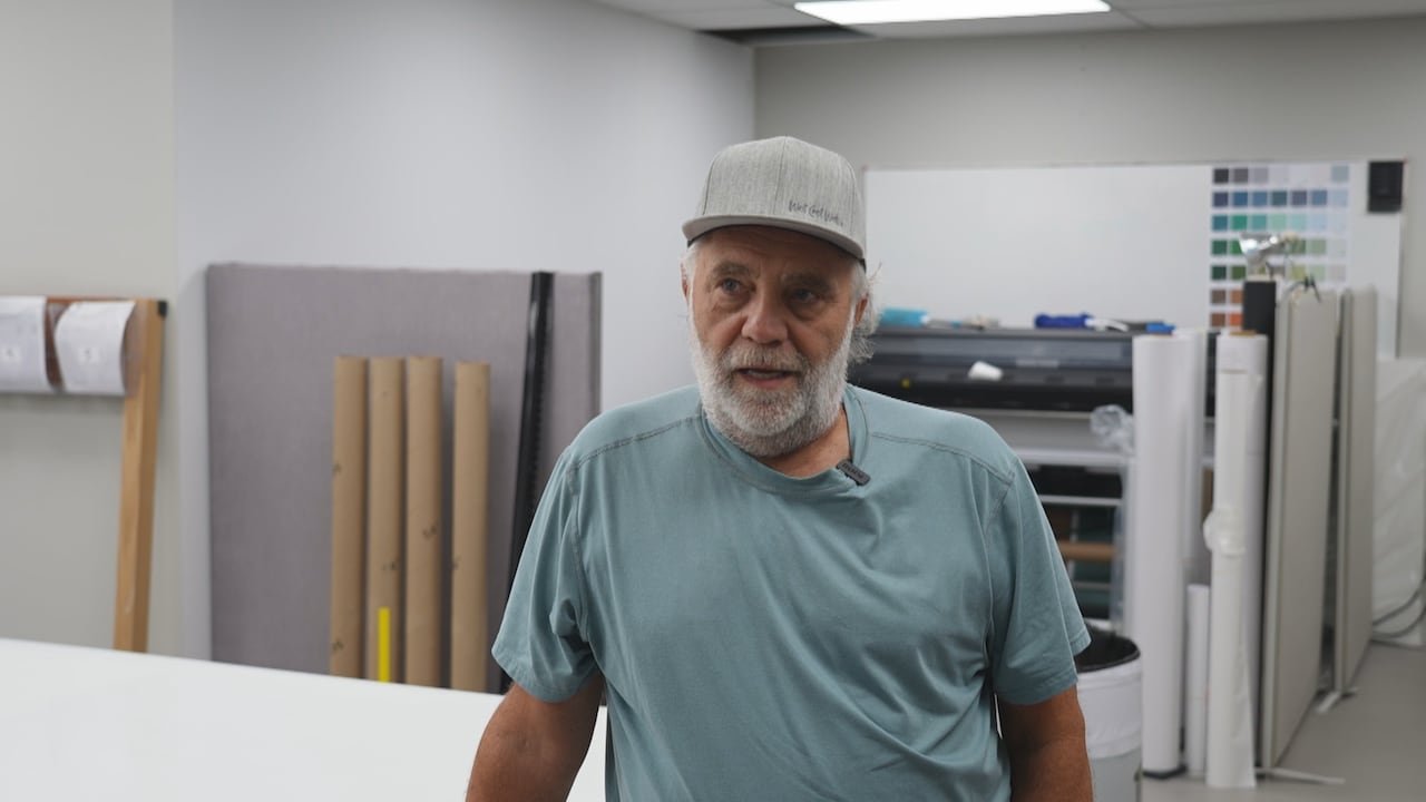 A man with a beard and a baseball cap surrounded by rolls of wallpaper.