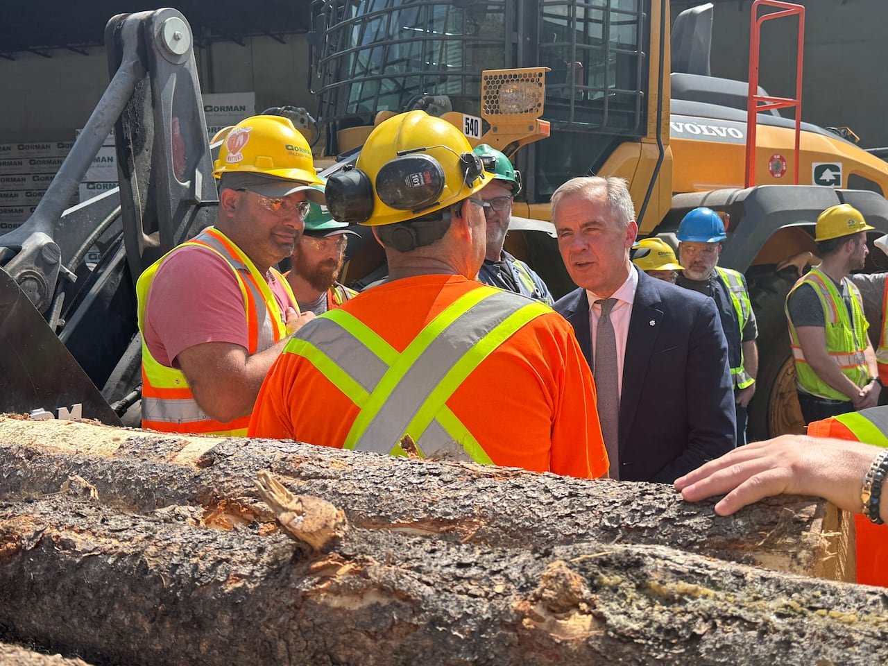 A man in a suit addressing mill workers wearing hard hats surrounded by logs.