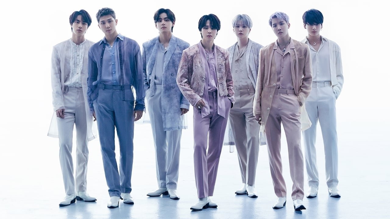 7 men in pastel-coloured outfits stand in a line against a white backdrop, with long shadows extending in front of them.