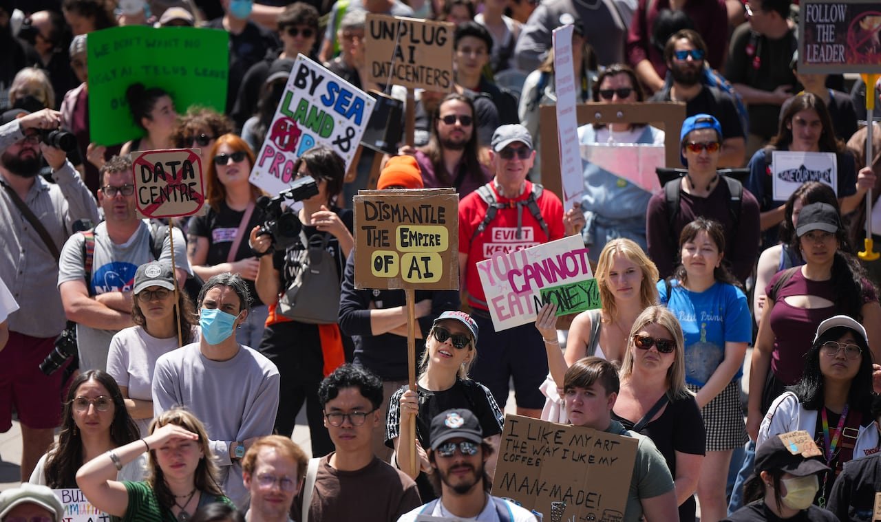 A large crowd of people fills the screen, many holding signs with messages against AI.