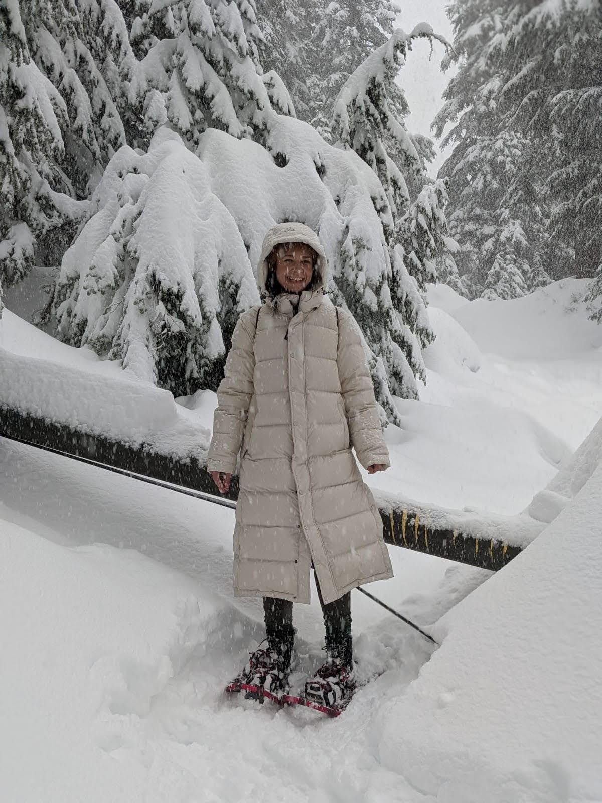 default-35 A woman in a white poofy jacket and snowshoes smiles as she poses on a snow-covered trail through tall pine trees.