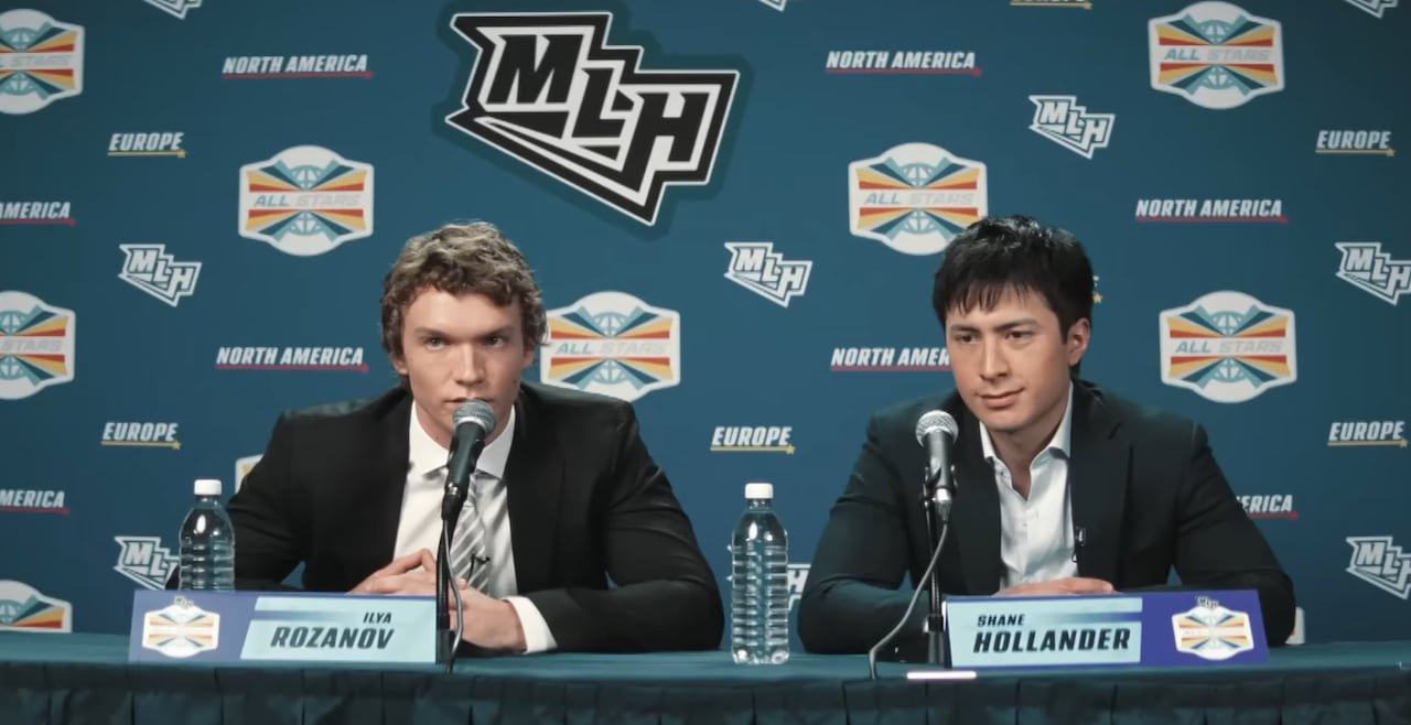 Two men sit down for a press conference. Both are wearing black blazers with white dress shirts underneath. Behind them is a series of logos