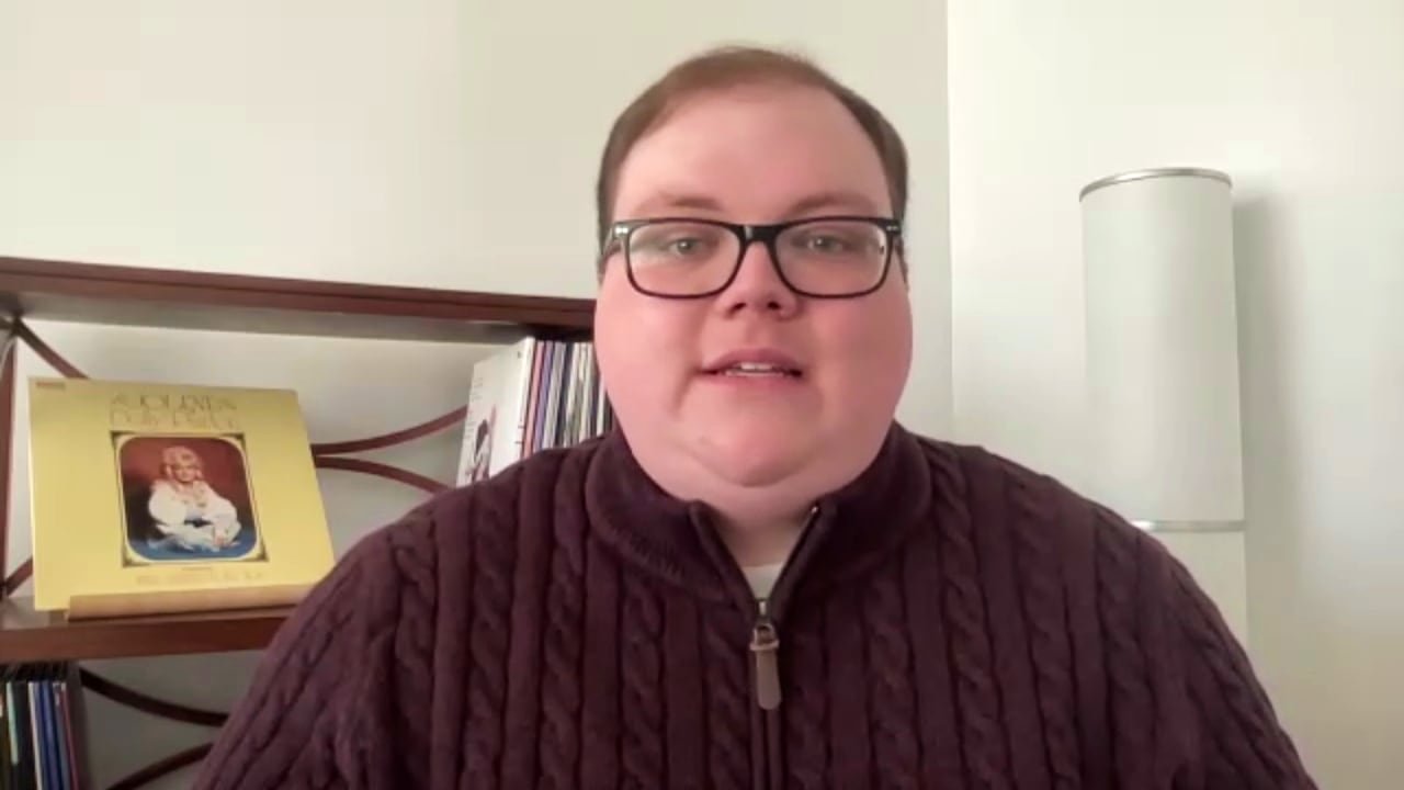 A man wearing a red sweater sits in an office. There's a Dolly Parton vinyl displayed on a shelf over his right shoulder.