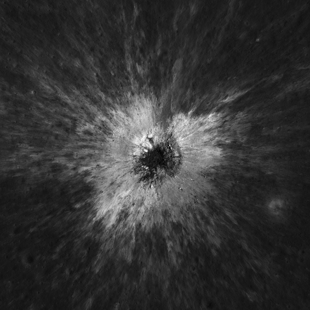An overhead view shows a crater on the moon with bright ejecta surrounding it.
